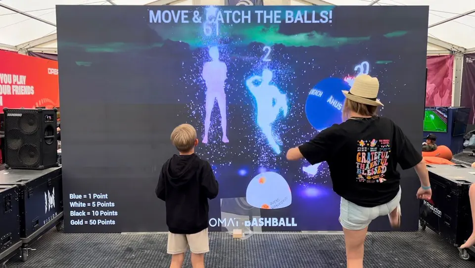 Zwei Kinder spielen Bashball vor einer großen LED-Wand, ihre Körperumrisse erscheinen als leuchtende Figuren im Spiel
